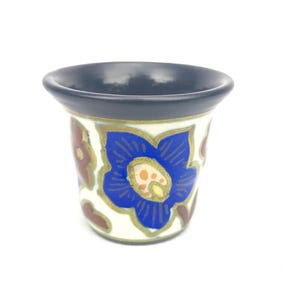 Może przedstawiać: Mały ceramiczny kubek z ciemnoniebieską obwódką i białym korpusem. Kubek jest ozdobiony dużym niebieskim kwiatem ze złotymi akcentami oraz brązowymi i złotymi wzorami kwiatowymi. Kubek ma rozszerzoną górę.