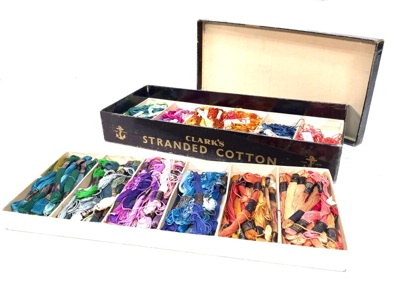 May include: A vintage box of Clark's Stranded Cotton embroidery floss. The box is black with gold lettering and an anchor design. The floss is arranged in rows of different colors, including green, blue, pink, and orange.