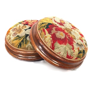 Peut inclure: Deux repose-pieds ronds avec des bases en bois brun foncé et des dessus rembourrés présentant un motif floral en point de tapisserie dans des tons de rouge, jaune, vert et crème. Chaque tabouret est orné d'une bordure en corde torsadée.