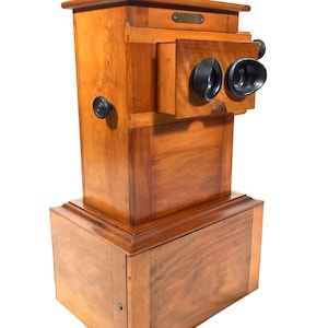 Op de afbeelding: Een vintage houten stereoscoop met een kijker met twee lenzen en een rechthoekige basis. Het antieke apparaat is gemaakt van gepolijst hout met zwarte knoppen en een klein metalen plaatje. Ontworpen voor het bekijken van 3D-afbeeldingen.