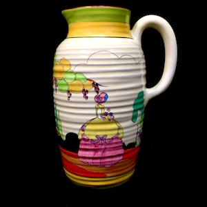 Könnte beinhalten: Vintage Keramikkrug mit weißem, geripptem Körper und gebogenem Griff. Der Krug ist mit einer farbenfrohen Illustration einer Frau in gelbem Kleid und rosa Rock mit grünen und lila Trauben verziert. Der Rand ist grün und gelb.