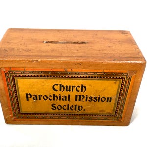 Könnte beinhalten: Eine Vintage-Holzkiste mit einem Schlitz oben für Spenden. Die Kiste hat eine rechteckige Form und ein dekoratives Etikett mit dem Text "Church Parochial Mission Society" in schwarzer Schrift auf gelbem Grund.