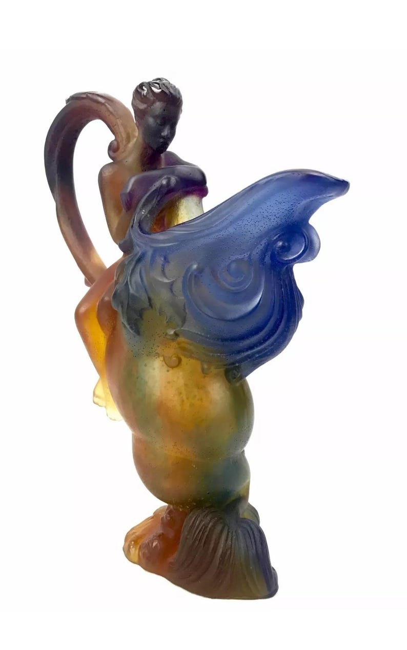 May include: A glass sculpture of a woman riding a winged horse. The sculpture is made of iridescent glass with shades of blue, green, and brown.