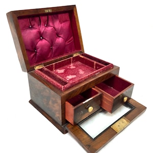 Op de afbeelding: Een houten juwelendoos met een donkerbruine wortelhouten afwerking, voorzien van een open deksel met dieprode satijnen voering, een uitneembaar rood fluwelen dienblad en twee kleine lades met goudkleurige knoppen en een glazen voorpaneel.