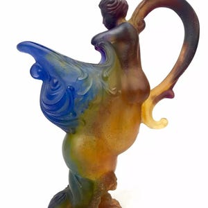 May include: A multi-colored glass pitcher with a figure of a woman riding a seahorse. The pitcher is made of iridescent glass with shades of blue, green, yellow, and brown.