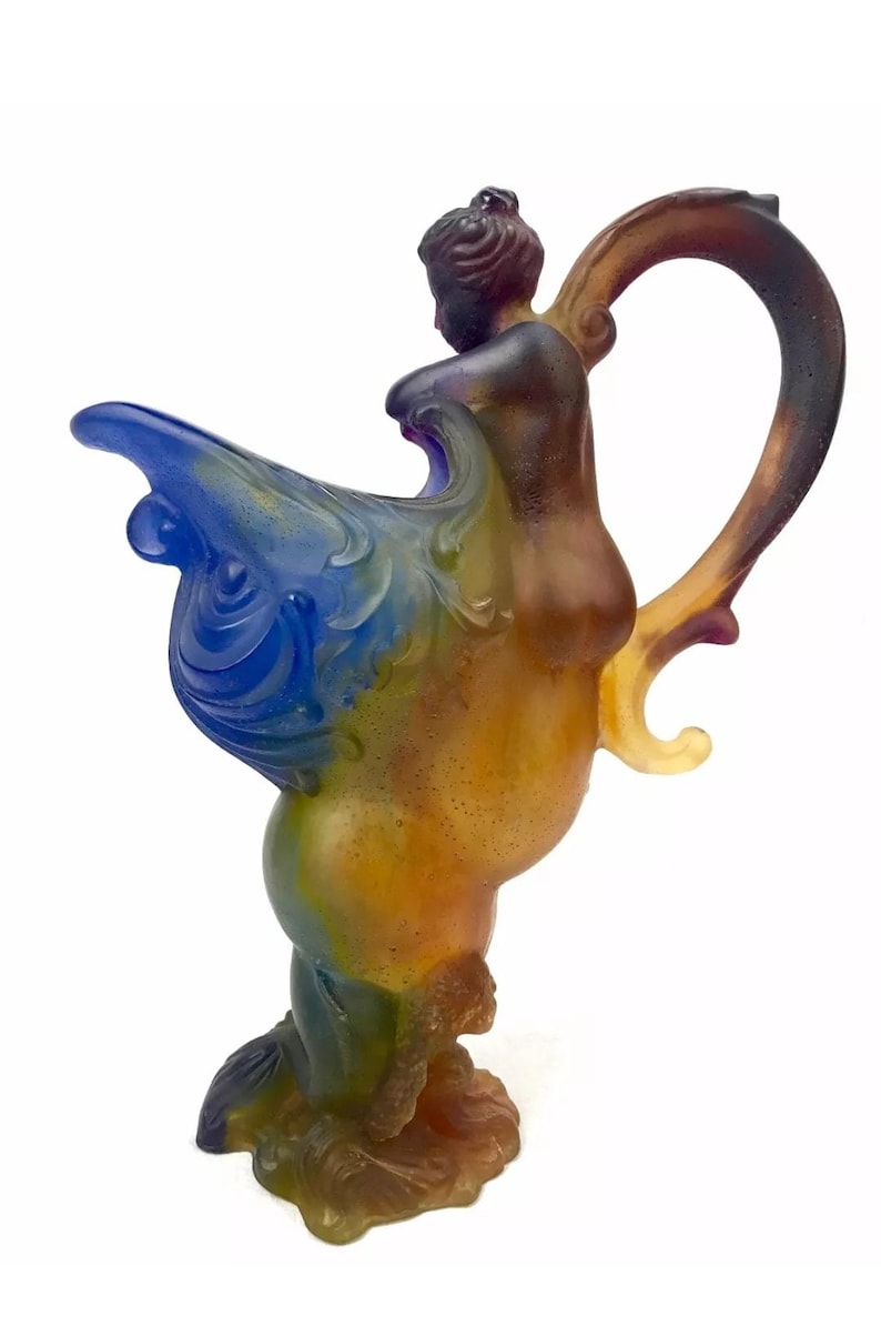 May include: A multi-colored glass pitcher with a figure of a woman riding a seahorse. The pitcher is made of iridescent glass with shades of blue, green, yellow, and brown.