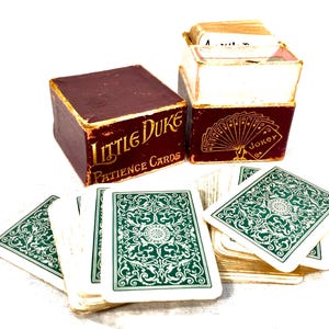 Può includere: Un mazzo di carte vintage "Little Duke Patience Cards" con una scatola bordeaux e scritte dorate. Il mazzo include carte da gioco con disegni verdi e bianchi e una scatola con un'illustrazione della carta Joker.