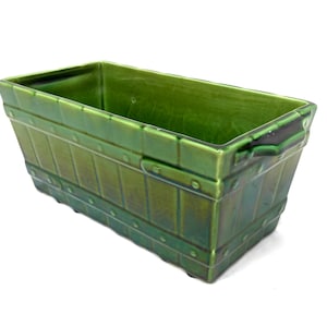 May include: A rectangular, green ceramic planter with a textured, paneled design. The planter has small, round rivet details and integrated handles. The glossy finish gives it a vintage look. Suitable for indoor or outdoor use.