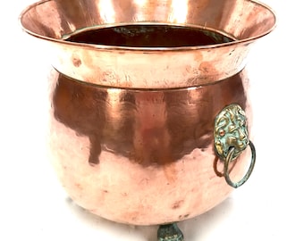 May include: A copper-colored metal pot with a wide rim and a rounded body. The pot features a decorative lion's head handle and rests on three small, ornate feet. The pot's surface has a polished, reflective finish.
