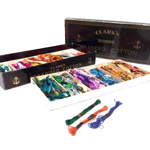 Op de afbeelding: Een houten doos gevuld met kleurrijke spoelen garen. De doos is gelabeld "Clark's Anchor Stranded Cotton".