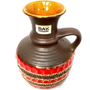 May include: A brown ceramic vase with a handle and a round body. The vase has a red and gold patterned design around the base and a brown top. The interior is a light brown color. The label on the vase reads "BAY KERAMIK".