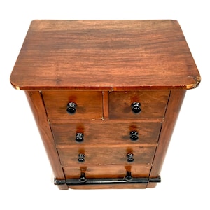 Può includere: Un piccolo comò in legno con cinque cassetti e pomelli neri. Il comò è in legno scuro e ha un design semplice.
