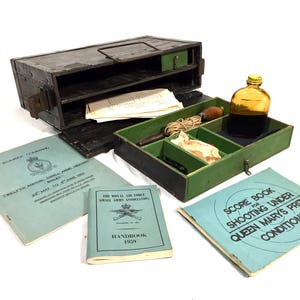 Pode incluir: Uma caixa de madeira com um interior verde que contém uma garrafa, uma corda e um livro intitulado "Score Book for Shooting Under Queen Mary's Prize Conditions".