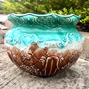 Può includere: Un vaso in ceramica caratterizzato da una sfumatura di colore che passa da una base marrone scuro a un bordo turchese vivace. L'esterno è decorato con un motivo a reticolo in rilievo, motivi floreali impressi e dettagli a volute.