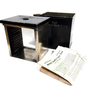 May include: An antique black metal Watkins Daylight Time Tank with a square base and a separate rectangular container. The tank has a series of internal slots. A folded instruction sheet is included.