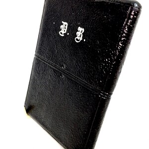 May include: A rectangular black wallet with a textured surface and a horizontal stitched seam across the middle. The front features the white embroidered initials A. B. in a decorative gothic script font, with a glossy finish on the material.