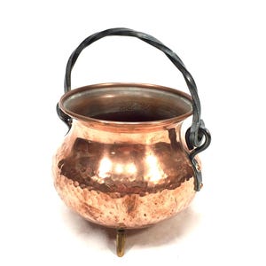 Peut inclure: Un chaudron en cuivre martelé de forme arrondie avec une large ouverture et un intérieur lisse. Le récipient repose sur trois petits pieds et possède une anse en métal torsadé sombre fixée sur les côtés par des boucles circulaires.