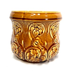 May include: A glazed ceramic planter with a glossy amber-brown finish, featuring a raised relief pattern of stylized flowers and foliage wrapping around the exterior. The vessel has a wide, smooth rim and a rounded, tapered body shape.