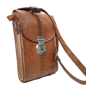 Könnte beinhalten: Eine vertikale braune Ledertasche mit strukturierter rechteckiger Form, einem Überschlag und einem silberfarbenen Metallverschluss. Die Tasche verfügt über einen langen, verstellbaren Schulterriemen aus Leder mit sichtbaren Nähten.