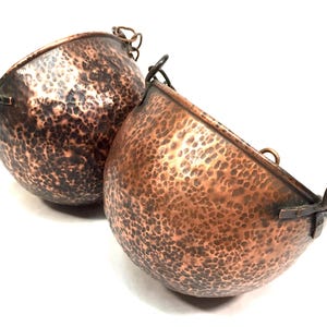 Peut inclure: Deux bols en cuivre avec une texture martelée et des taches sombres. Chaque bol a une forme arrondie avec une poignée de chaque côté. Les bols sont posés sur un fond blanc.