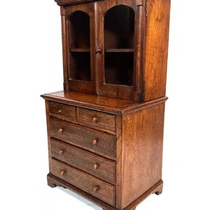 Può includere: Un antico mobile in legno scuro con una sezione superiore in vetro e cinque cassetti sotto. Il mobile ha una parte superiore decorativa e un design semplice.
