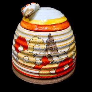 May include: A ceramic honey pot shaped like a beehive with a bee on top. The pot is decorated with orange, yellow, and white stripes and flowers.
