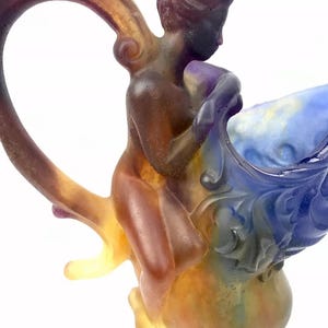 May include: A glass sculpture of a woman riding a large, colorful, glass shell. The woman is nude and has a bun in her hair. The shell is blue, purple, and white. The sculpture is made of glass and has a textured surface.