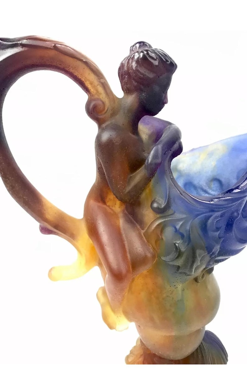 May include: A glass sculpture of a woman riding a large, colorful, glass shell. The woman is nude and has a bun in her hair. The shell is blue, purple, and white. The sculpture is made of glass and has a textured surface.