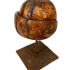 May include: A wooden hat block with a rounded shape, made of multiple pieces of wood, mounted on a rusty metal stand.