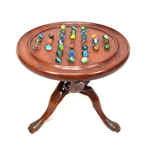 May include: A vintage wooden marble game table with an oval playing surface. The table features a dark brown finish and is supported by three carved legs. Colorful glass marbles are arranged on the playing surface, ready for play.