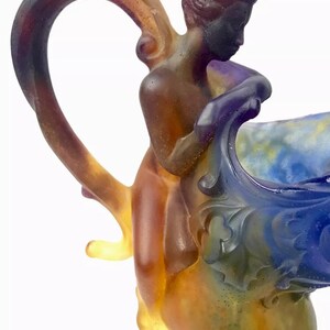 May include: A glass pitcher with a colorful, iridescent finish. The pitcher features a sculpted figure of a woman holding onto the rim. The handle is shaped like a vine and is also iridescent.