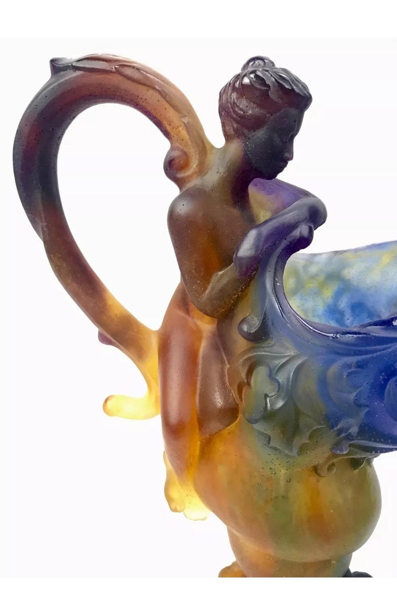 May include: A glass pitcher with a colorful, iridescent finish. The pitcher features a sculpted figure of a woman holding onto the rim. The handle is shaped like a vine and is also iridescent.