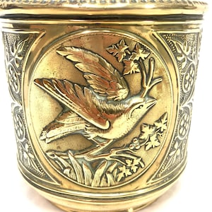 May include: A detailed brass container with a bird and floral relief design. The bird is perched on a branch, holding a twig in its beak. The container has a decorative rim and side panels with floral patterns.