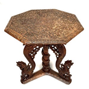 May include: An octagonal wooden table with a dark brown finish, featuring an intricate floral relief carving on the top surface. The base consists of three carved dragon-shaped legs and a central pillar with a coiled serpent design.