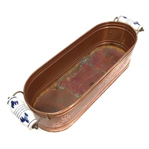 Puede incluir: Una cubeta ovalada de cobre con textura exterior martillada y una pátina interior desgastada. El recipiente incluye dos asas laterales de cerámica blanca con motivos florales azules, unidas al cuerpo de cobre con herrajes metálicos.