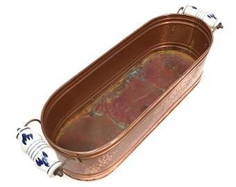 May include: An oval copper basin featuring a hammered exterior texture and a weathered interior patina. The vessel includes two side handles made of white ceramic with blue floral patterns, attached to the copper body with metal hardware.