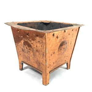 May include: A square, copper-coloured planter with a weathered appearance. The planter has a flared design, decorative circular accents, and is supported by four small legs. The edges are slightly bent, adding to its vintage aesthetic.