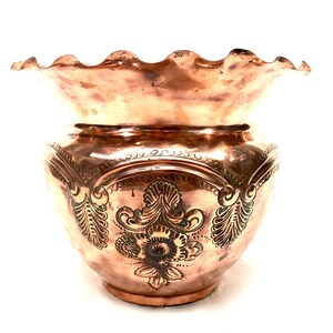 May include: A copper-coloured decorative bowl with a ruffled rim. The bowl features ornate, embossed floral and leaf patterns around the body. The metal has a polished finish, reflecting light. The bowl is likely for decorative purposes.