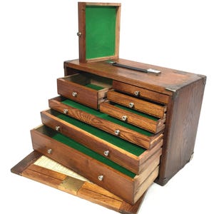 May include: A wooden machinist chest with a dark brown finish and metal corner reinforcements. The chest features multiple drawers with green felt linings, a top compartment with a green felt lid, and a drop-down front panel revealing a paper chart.