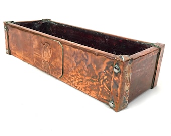 May include: A long, rectangular copper planter with a hammered finish. The planter has a raised, rectangular design on the front with a floral motif.