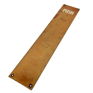 Peut inclure: Plaque de porte rectangulaire en laiton vieilli avec le mot "PUSH" en lettres blanches. La plaque comporte quatre trous de vis et une surface usée et texturée, suggérant une utilisation vintage.