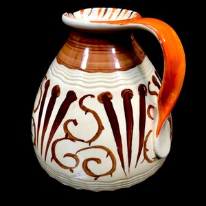 May include: A white ceramic jug with a brown and orange design. The jug has a wide, round body and a short, curved handle. The design features brown swirls and lines, as well as a few brown dots.