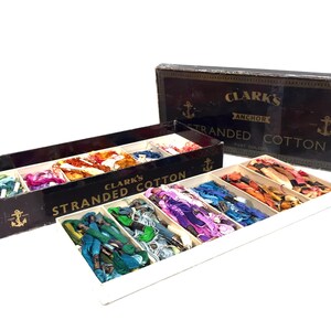 May include: A box of Clark's Anchor Stranded Cotton embroidery floss in a variety of colors. The box is open and the floss is arranged in rows. The box is black with gold lettering and an anchor logo.