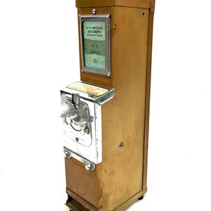 May include: A vintage mustard-colored metal U.S. postage stamp vending machine with a chrome coin mechanism. The display window reads U.S. POSTAGE STAMPS and 4-1c STAMPS 5c. A metal key is inserted into the top lock of the rectangular vertical unit.