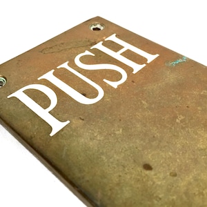 Op de afbeelding: Een rechthoekige messing deurplaat met het woord "PUSH" in witte hoofdletters. De plaat heeft twee schroefgaten en een verweerde, verouderde uitstraling, wat suggereert dat hij vintage of antiek is.