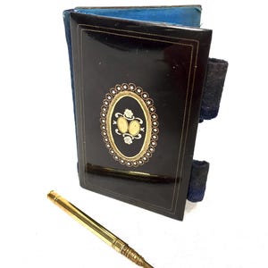 Op de afbeelding: Een zwart lederen notitieboekje met gouden en witte decoratieve accenten. Het notitieboekje heeft een blauwe fluwelen voering en wordt geleverd met een gouden potlood.