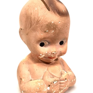May include: A vintage ceramic piggy bank shaped like a baby's head with a slot on top for coins. The baby has painted eyes, a red mouth, and is holding a heart in its hands.