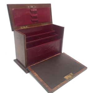 May include: A wooden desk organizer with a hinged lid and a fold-down writing surface. The interior is lined with red material and features compartments for storage. The organizer is made of dark wood and has brass hardware.
