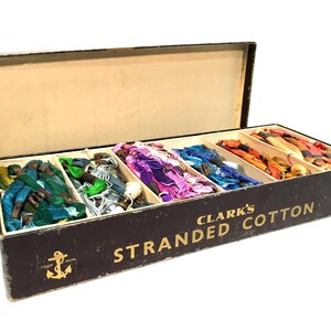 May include: A vintage box of Clark's Stranded Cotton embroidery floss. The box is black with gold lettering and an anchor logo. It contains multiple spools of colorful floss, including green, blue, purple, red, orange, and yellow.