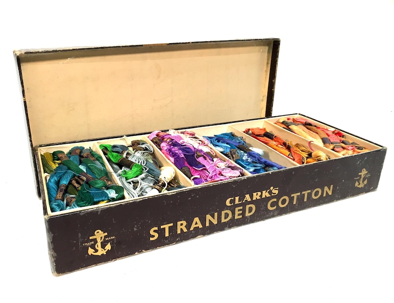 May include: A vintage box of Clark's Stranded Cotton embroidery floss. The box is black with gold lettering and an anchor logo. It contains multiple spools of colorful floss, including green, blue, purple, red, orange, and yellow.